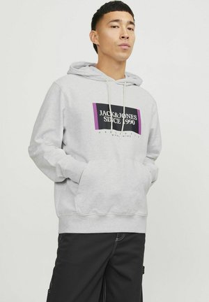 Sivi dukserica s kapuljačom koja ima prednji džep i vezicu. Sadrži središnji grafički prikaz u crnoj i ljubičastoj boji s natpisom "JACK & JONES SINCE 1990."