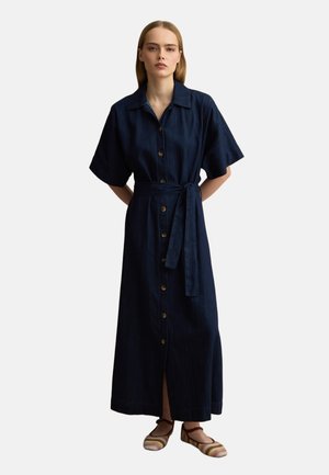 Vrouw staand met handen achter haar rug, draagt een lange marineblauwe jurk met knopen, korte mouwen en een gestrikte tailleband, plus gestreepte platte schoenen.