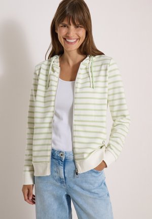 Femme souriante portant un sweat à capuche zippé rayé vert clair et blanc avec cordons, un débardeur blanc et un jean bleu clair, main dans la poche.
