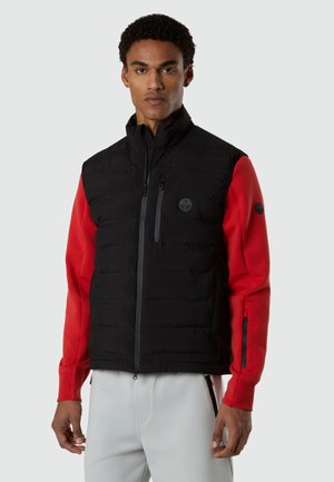 Uomo che indossa un gilet trapuntato nero sopra una maglia rossa a maniche lunghe e pantaloni grigio chiaro, in piedi contro uno sfondo chiaro e uniforme.