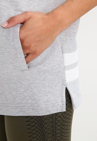 Šedé sportovní tričko s boční kapsou, má kontrastní bílý síťovaný pruh na pravé straně a zakřivený spodní lem pro zdůraznění detailů.