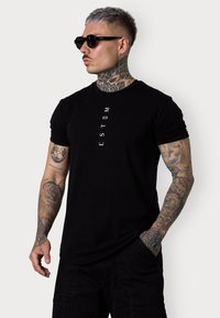 Schwarzes T-Shirt mit vertikalem weißen Text "ESTEM" auf der Vorderseite, kurze Ärmel, getragen mit schwarzen Sonnenbrillen, mit tätowierten Armen und einem schlichten Hintergrund.