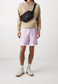 Sweat à capuche beige, short lavande avec bordure blanche, et baskets de couleur claire avec semelle en gomme. Sac bandoulière noir avec zips et logo.