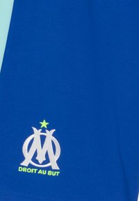Zils audums ar baltu savītu "OM" logo, virs kura ir neliels zaļš zvaigznīte, un zem logo ir dzeltenie uzraksti "DROIT AU BUT".