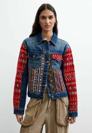 Donna con lunghi capelli scuri che indossa una giacca di jeans con motivi ricamati colorati sul davanti e maniche rosse decorate.