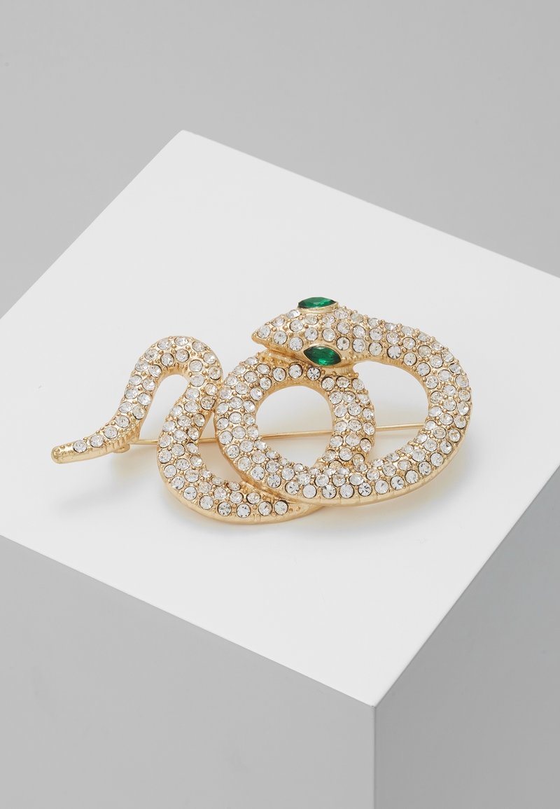 Broche en forme de serpent en or avec des strass clairs et deux yeux en pierres précieuses vertes, présentée sur une surface blanche avec un fond gris.