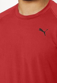 Rotes T-Shirt mit kurzen Ärmeln aus glattem Stoff, das ein schwarzes Puma-Logo auf der linken Brustseite und einen Rundhalsausschnitt aufweist.