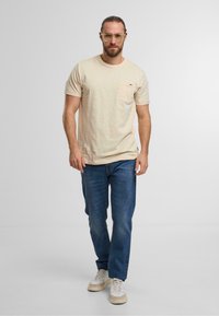 Beige t-shirt met korte mouwen en een zak, gedragen met blauwe jeans en beige sneakers. De stof lijkt zacht met een gestructureerde afwerking.