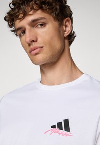 Hvid bomulds t-shirt med et sort logo med tre striber og pink tekst. Rund hals med en glat, blød tekstur.