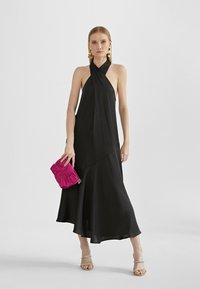 Black halter-neck dress with a flowing silhouette. Sheer fabric, asymmetrical hem. Model holds a textured pink clutch with a knot detail.