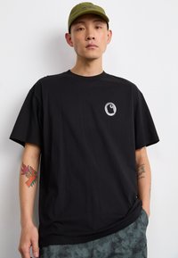 Camiseta de algodón negra con cuello redondo, que presenta un pequeño logo gris en el pecho. Combinada con pantalones cortos con estampado de camuflaje y una gorra verde.