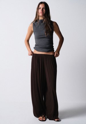 Jeune femme aux longs cheveux portant un haut gris sans manches et un pantalon noir ample, ajustant la taille du pantalon, debout sur un fond uni.