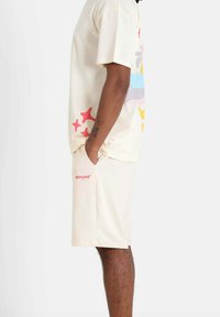 Beige cotton t-shirt with colorful graphic prints and matching beige shorts featuring a small red logo on the side. Side view displayed.