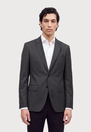 Un homme pose dans un costume gris anthracite à deux boutons, une chemise blanche et un pantalon foncé, sur un fond clair uni.