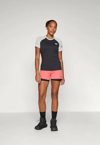 Črna in siva športna majica s kratkimi rokavi v kombinaciji z roza kratkimi hlačami. Model nosi črne nogavice in čevlje. Preprost, funkcionalen dizajn.