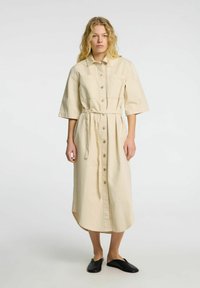 Beige denim skjortekjole med krave, korte ærmer, knaplukning foran og bindebælte; har lommer og en buet kant.