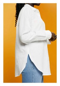 Chemise blanche à coupe décontractée, en tissu texturé, avec un col évasé, des manches longues et des fentes latérales. Associée à un jean bleu.