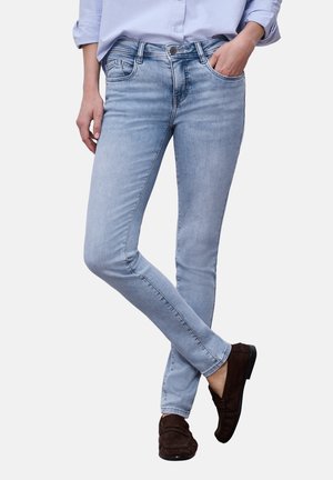 Person iført lyseblå slim-fit jeans parret med brune loafers, stående med det ene ben krydset over det andet.