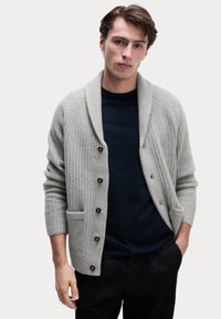 Cárdigan gris con canalé y cuello chal, botones negros y dos bolsillos frontales, superpuesto sobre una camisa azul marino, combinado con pantalones oscuros.