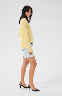 Suéter de punto amarillo claro, corte holgado, mangas largas. Falda de denim azul claro desgastada. Tacones negros de punta afilada con tiras detrás. Accesorios mínimos.