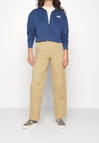 Felpa blu navy tagliata con doppie tasche frontali, pantaloni cargo beige con cuciture, e scarpe slip-on a scacchi bianco e nero.