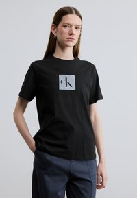 Μαύρο βαμβακερό μπλουζάκι με σχέδιο λογότυπου σε σχήμα τετραγώνου με τα γράμματα "cK" σε μεταλλικό ασημί. Λείο ύφασμα, κοντά μανίκια, λαιμόκοψη τύπου στρογγυλή.