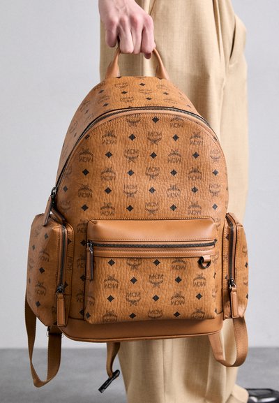 Zaino in pelle marrone con logo MCM impresso, motivo a diamante, forma tondeggiante, tasche laterali doppie e scomparto anteriore con zip.
