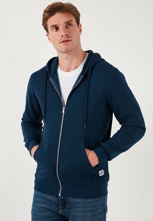 Marineblå zip-up hoodie i bomuldsblanding, med snoretræk i hætten, frontlommer og et lille logo-klistermærke på kanten.