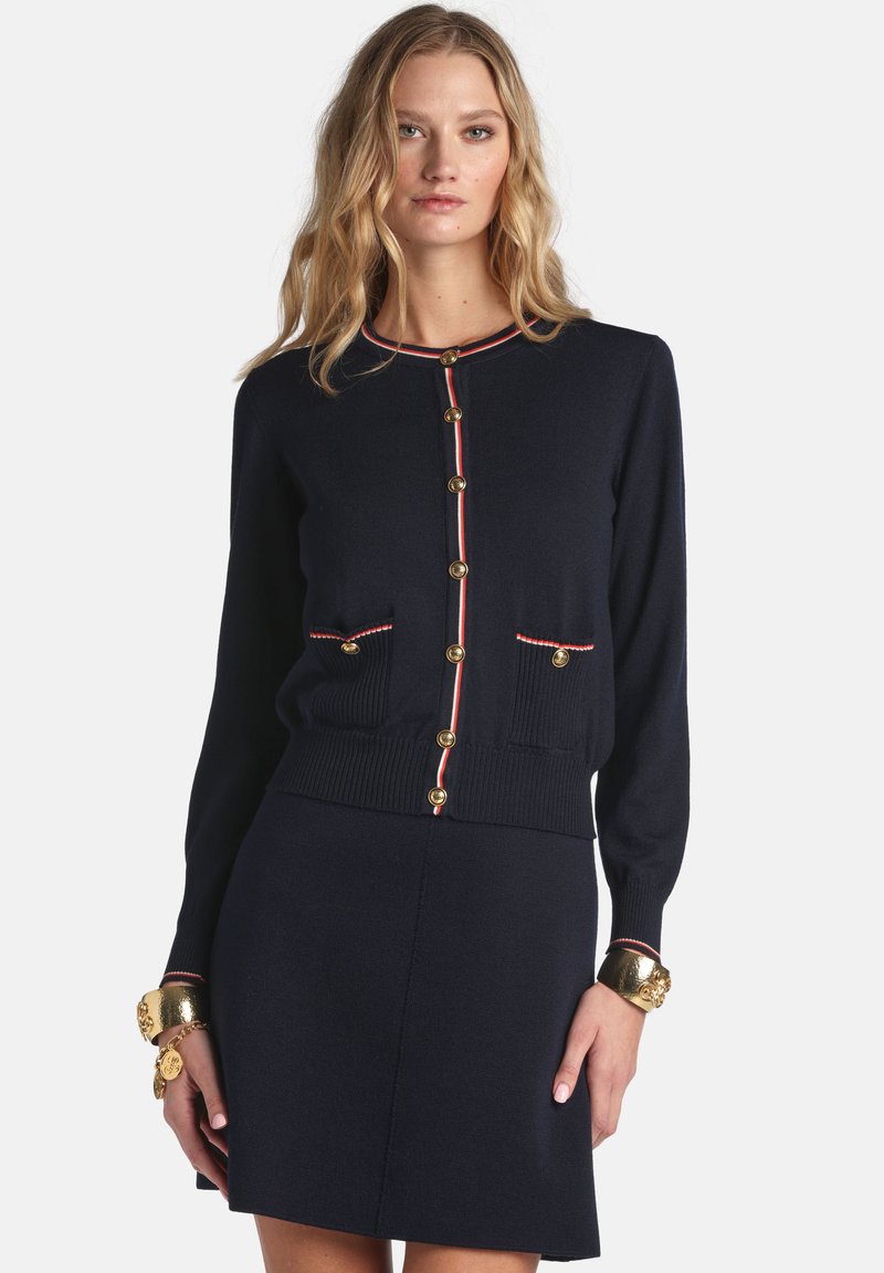 Marineblauer Cardigan mit goldenen Knöpfen und zwei Fronttaschen, mit rotem und weißem Besatz entlang des Halsausschnitts und der Bündchen. Weiche Strickstruktur.