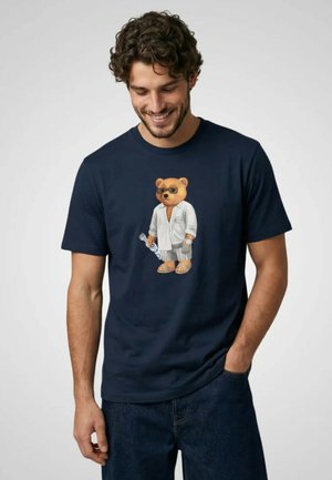 Uomo che indossa una t-shirt blu navy con un orso stampato con occhiali da sole, camicia bianca, pantaloncini a righe e sandali che tiene una bottiglia.
