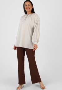 Blusa lunga beige chiaro con scollo arricciato, abbinata a pantaloni marroni aderenti e scarpe aperte di colore chiaro.