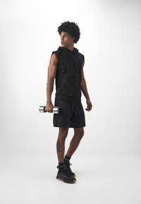 Fekete ujjatlan kapucnis felső elöl zsebbel, hozzá illő sorttal, ezüst súlyzót tartva. Fekete cipő és zokni van rajta. Egyszínű háttér.