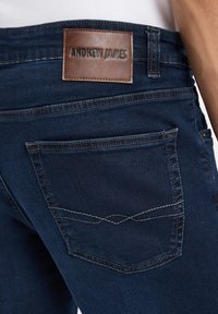 Dunkelblaue Jeans bestehen aus einem braunen Lederpatch mit der Prägung "ANDREW JAMES". Die Tasche weist einen kontrastierenden Steppnahtdetail auf.