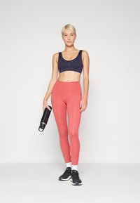 Svart sports-bh, rosa høyt liv leggings med sømdetaljer, hvite sokker, svarte treningssko, og en svart vannflaske i hånden.