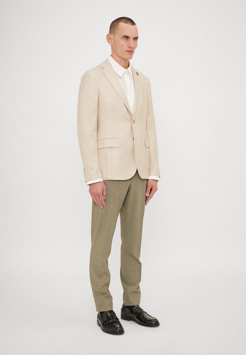 Blazer beige chiaro con un design in lino testurizzato, abbinato a pantaloni verde oliva. Scarpe nere lucide completano l'outfit.