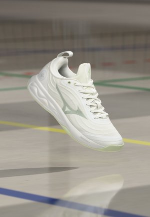 Weißer Sportschuh mit silbernem Logo, Schnürsenkeln und hellgrüner Sohle, der über einem Hallenboden mit farbigen Spielfeldlinien schwebt.