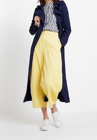 Marinblå trenchcoat, randig skjorta med knappar och gula vida byxor. Vita sneakers fulländar outfiten med en avslappnad passform och textur.