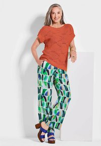 Oranger Strickpullover mit kurzen Ärmeln, gemusterte grüne und blaue weite Hosen und blaue Absatzsandalen. Glatte Texturen und leichtes Material.