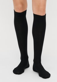 Calze nere fino al ginocchio con una texture a costine, dal finish morbido e con punta arrotondata. Il risvolto superiore è liscio ed elastico.