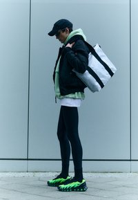 Svart pufferjakke over en mintgrønn hettegenser, svarte tights og grønne sneakers med detaljer. Bærer en hvit tote-bag med svarte striper.
