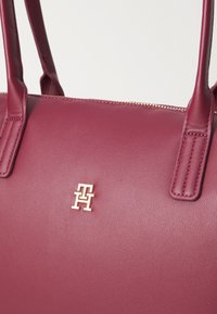 Sac à main en cuir bourgogne avec deux poignées, accent logo doré, texture lisse et fermeture éclair sur le bord supérieur.