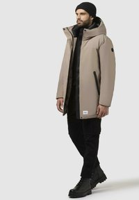 Beige polstret parka med høj krave, lynlås og knaplukning, sidelommer og en sort accent. Bæres med sorte bukser og støvler.