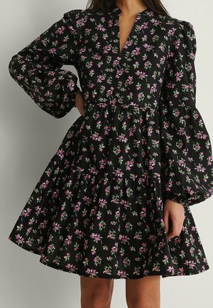 Robe noire avec manches longues bouffantes et jupe à étages, ornée de petits motifs floraux roses et verts, portée par une personne debout.