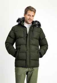 Olijfgroene pufferjack met een nepbont capuchon, voorzakken met ritssluiting en een gequilt textuurontwerp. Gecombineerd met olijfgroene broek.