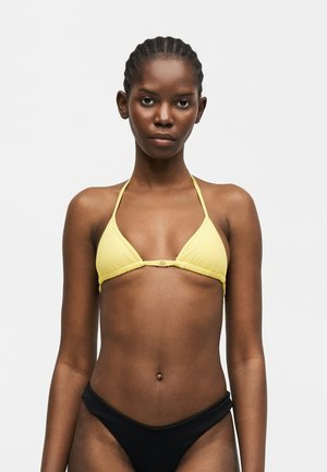 Žena s pletenicom u kosi koja nosi žuti trokutasti gornji dio bikinija i crni donji dio bikinija, stoji na jednobojnoj svijetloj pozadini.