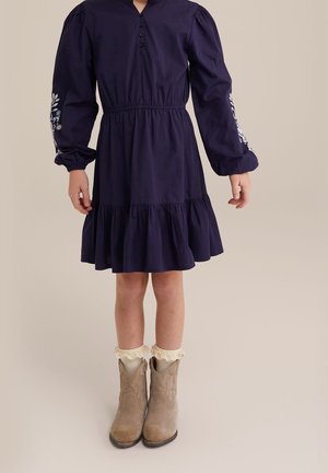 Marineblaues Baumwollkleid mit langen Puffärmeln, geraffter Taille und Rüschensaum. Verziert mit weißer Blumenstickerei an den Ärmeln. Braune Stiefeletten.