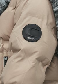 Μπεζ επενδυμένο μπουφάν με υφή και μαύρο στρογγυλό λογότυπο στο μανίκι, διαθέτει κλείσιμο με φερμουάρ και λεία υφή υφάσματος.