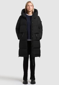 Cappotto piumino nero con cappuccio, caratterizzato da un design trapuntato, tasche con zip e un taglio a lunghezza media, abbinato a leggings scuri e stivali.