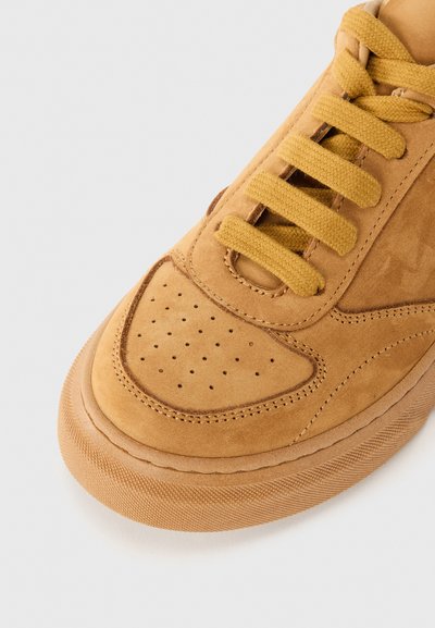 Sneaker en daim marron clair avec un orteil perforé, des lacets plats jaunes et une semelle en caoutchouc texturée. Présente des coutures lisses et un col rembourré.