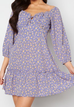 Kvinde iført en lilla blomsterprikket off-shoulder kjole med lange ærmer, flæsekant og bindedetalje ved halsudskæringen, der holder kjolen ud.